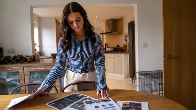 Sasha-Jay observa las múltiples copias en tamaño A4 de las cuentas falsas que están sobre la mesa de su cocina. Con la mano derecha sostiene una pila de imágenes, mientras que con la izquierda toca una de ellas.