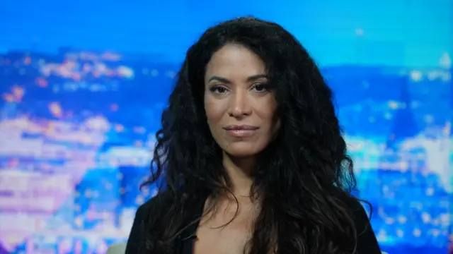 Lisa Phillips, con el pelo negro ondulado, mira al frente. Lleva una blusa negra. Al fondo se divisa un paisaje urbano azul y morado.