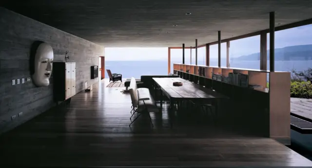 Interior oscuro con techo de hormigón y un comedor abierto hacia el mar mediante grandes ventanales.