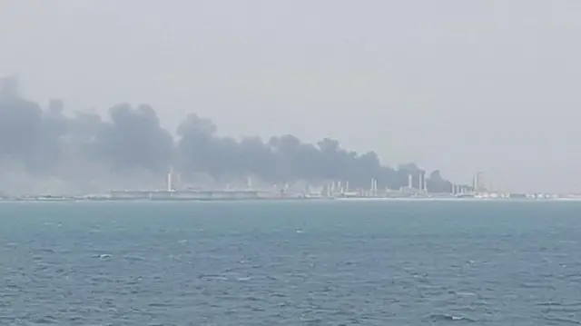 Columnas de humo se elevan desde la derecha de la foto sobre edificios a lo largo de la costa. El mar azul oscuro está en primer plano.