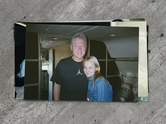 Bill Clinton, un hombre mayor con el pelo corto y gris, está de pie junto a una joven, Chauntae Davies. Clinton lleva una camiseta negra de manga corta con el logo de Air Jordan. Davies viste una chaqueta vaquera de color claro. Se encuentran dentro de un avión.
