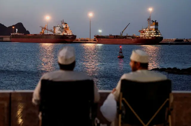 Dos buques cisterna permanecen anclados el 9 de marzo de 2026. En primer plano, dos hombres con túnicas blancas están sentados en sillas, contemplando el canal y los barcos.