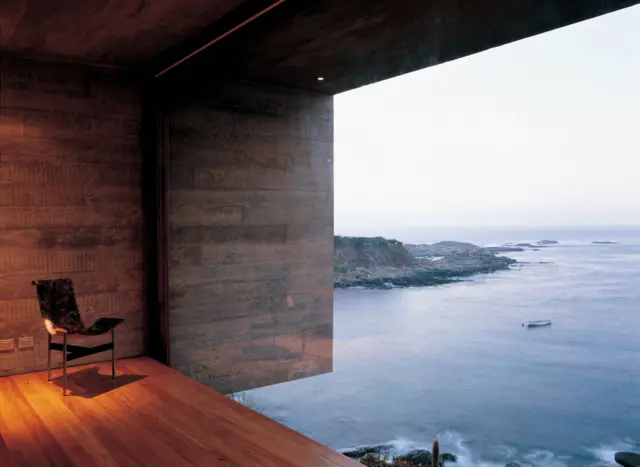 Espacio interior con muros de hormigón y piso de madera que se abre mediante un gran ventanal hacia una vista costera amplia.