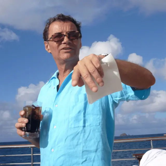 Un hombre con camisa azul claro de manga corta y gafas de sol, sostiene una bebida en una mano y un papel en la otra.