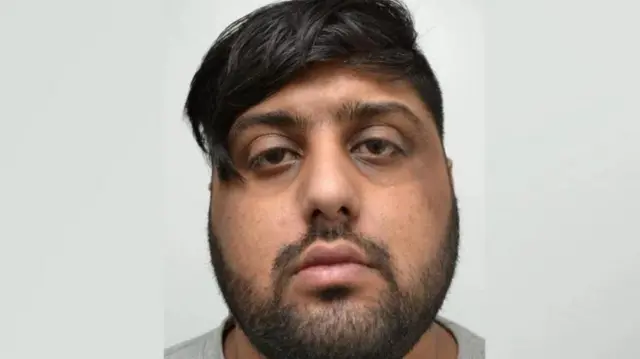 Un hombre de cabello y barba negros mira hacia la cámara. Lleva puesta una prenda superior gris y se encuentra frente a un fondo blanco liso.