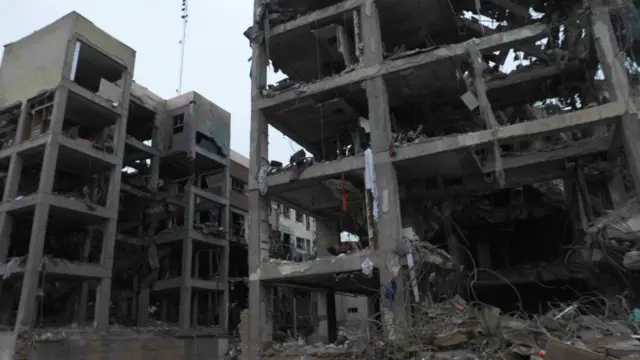 Dos edificios destruidos, de cuatro a cinco pisos de altura, con las ventanas y las paredes reventadas. Permanecen en pie algunos pilares y losas de hormigón. Hay algunos trozos de tela colgando de la estructura y metal retorcido en el suelo.