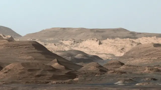 La imagen muestra las regiones más altas del Monte Sharp en Marte. En primer plano se aprecian algunas zonas más oscuras con pequeños montículos, y a la izquierda se observa una pendiente ligeramente más pronunciada. En el centro de la foto hay algunas zonas rocosas más claras y de aspecto escarpado. A lo lejos se ven colinas más grandes, de formas más redondeadas y con pendientes suaves. El cielo, que se divisa en la parte superior, es de color gris oscuro.