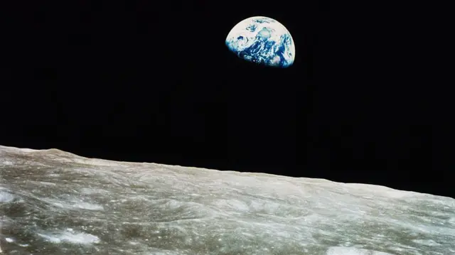 En primer plano se ve una pequeña sección de la superficie lunar plateada, con una vista parcial de la Tierra como una bola azul con remolinos blancos que "se elevan" en un "cielo" negro azabache.