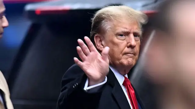 Donald Trump saluda de lado. Viste traje oscuro y corbata roja.