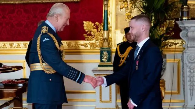 El rey Carlos, vistiendo un uniforme de la RAF, sonríe a Nathan Newby —un hombre con traje azul oscuro— mientras le estrecha la mano. Al fondo se observa a otro hombre con uniforme militar, de pie en una estancia de aspecto majestuoso, con decoraciones doradas y una chimenea de mármol tallado.