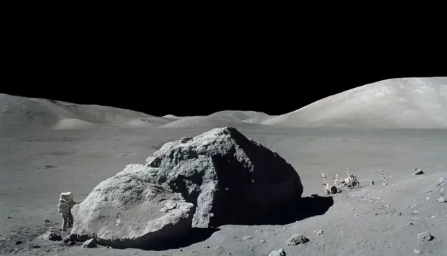 Una foto en blanco y negro de una enorme roca en la superficie rocosa de la Luna y un astronauta del lado izquierdo.