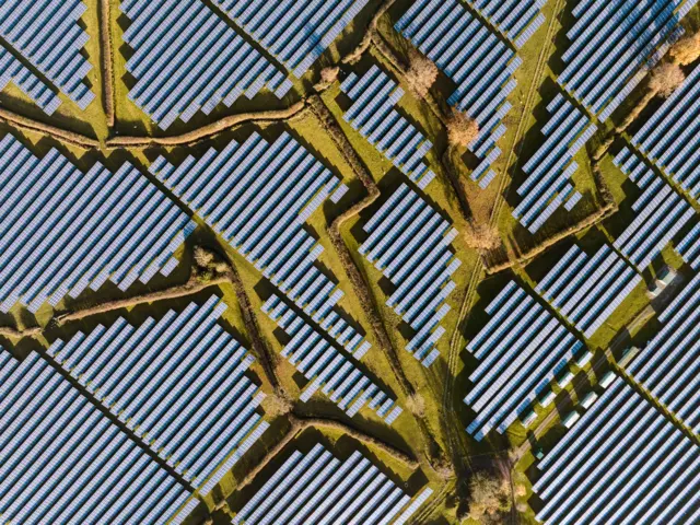 Una toma aérea muestra hileras de paneles solares dispuestos en patrones geométricos, atravesadas por estrechos senderos o canales. Las formas repetitivas crean un paisaje con textura, similar a una cuadrícula.