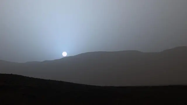 Una foto tomada desde la superficie de Marte del Sol poniéndose justo sobre el horizonte montañoso. El cielo tiene un tono azul cerca del Sol y se ve gris a mayor distancia.