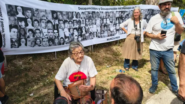 Una mujer da una entrevista mientras atrás hay una pancarta con rostros de personas desaparecidas
