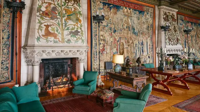 Sala con sillones verdes en torno a una estufa a leña, mesas de madera y pisos de madera con alfombras. Enormes tapices cubren las paredes.