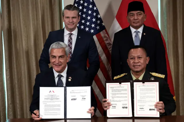 El secretario de Defensa de EE.UU., Pete Hegseth, y el ministro de Defensa de Indonesia, Sjafrie Sjamsoeddin, están de pie detrás de un escritorio, mientras sus colegas sentados sostienen en alto un memorando de entendimiento.