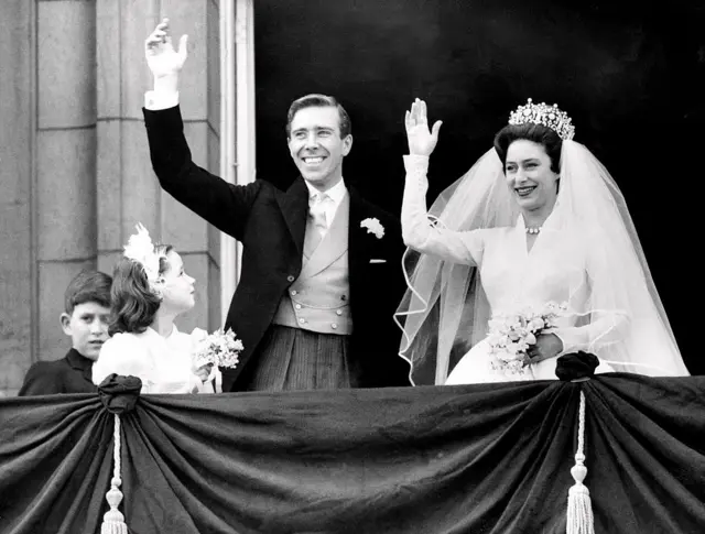 Una pareja de recién casados, él de sacoleva y ella de blanco con velo y una corono, saludan desde un balcón con los brazos en alto.