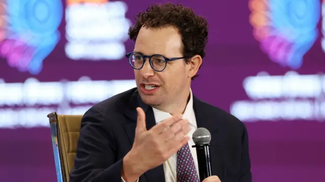 El director ejecutivo de Anthropic, Dario Amodei, se dirige a los asistentes en la Cumbre de Impacto de la IA, en Nueva Delhi, India. Viste gafas, un traje oscuro y una corbata morada, sentado frente a un fondo morado. Sostiene un micrófono mientras se dirige a alguien invisible.