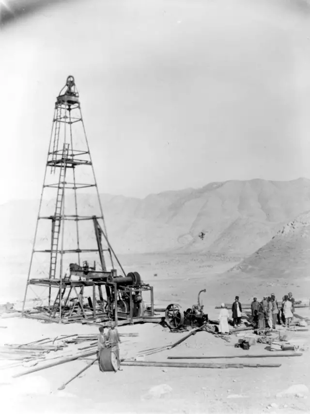 Una foto en blanco y negro muestra la construcción de una primitiva torre de extracción de petróleo en una región desértica con unas diez personas alrededor
