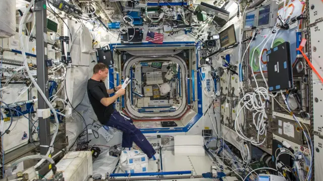 Wiseman flota en la ingravidez dentro de un módulo de la estación espacialm rodeado de cables y paneles de sistemas. El astronauta lee un manual mientras sostiene un lápiz entre los dientes