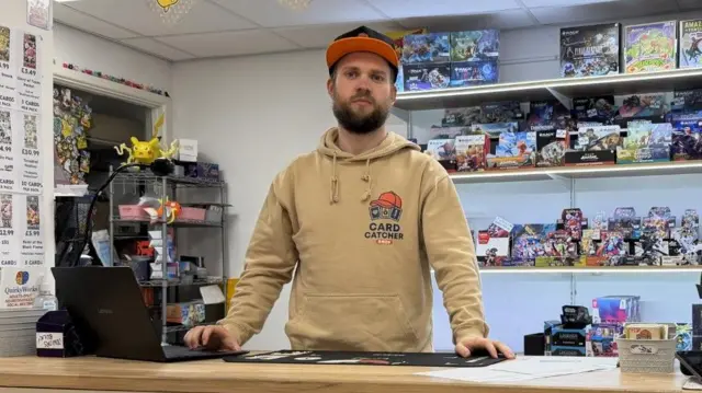 Sam Jackway, propietario de la tienda Card Catcher Shop en Bristol, está de pie junto al mostrador, con cartas Pokémon y otros objetos de colección en los estantes detrás de él.