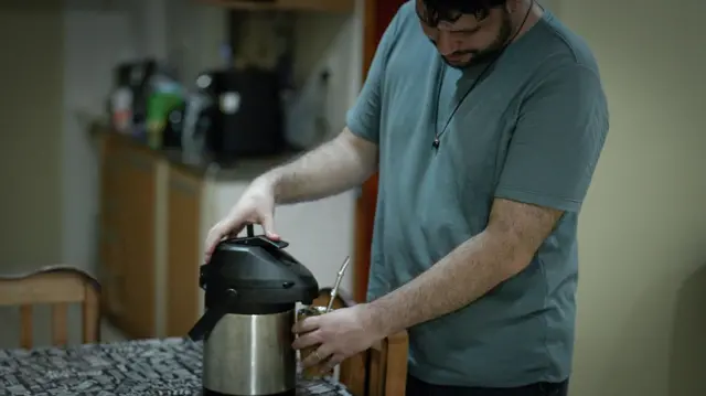 Un hombre con playera verde, prepara en una cocina su infusión de yerba mate con agua de un termo.