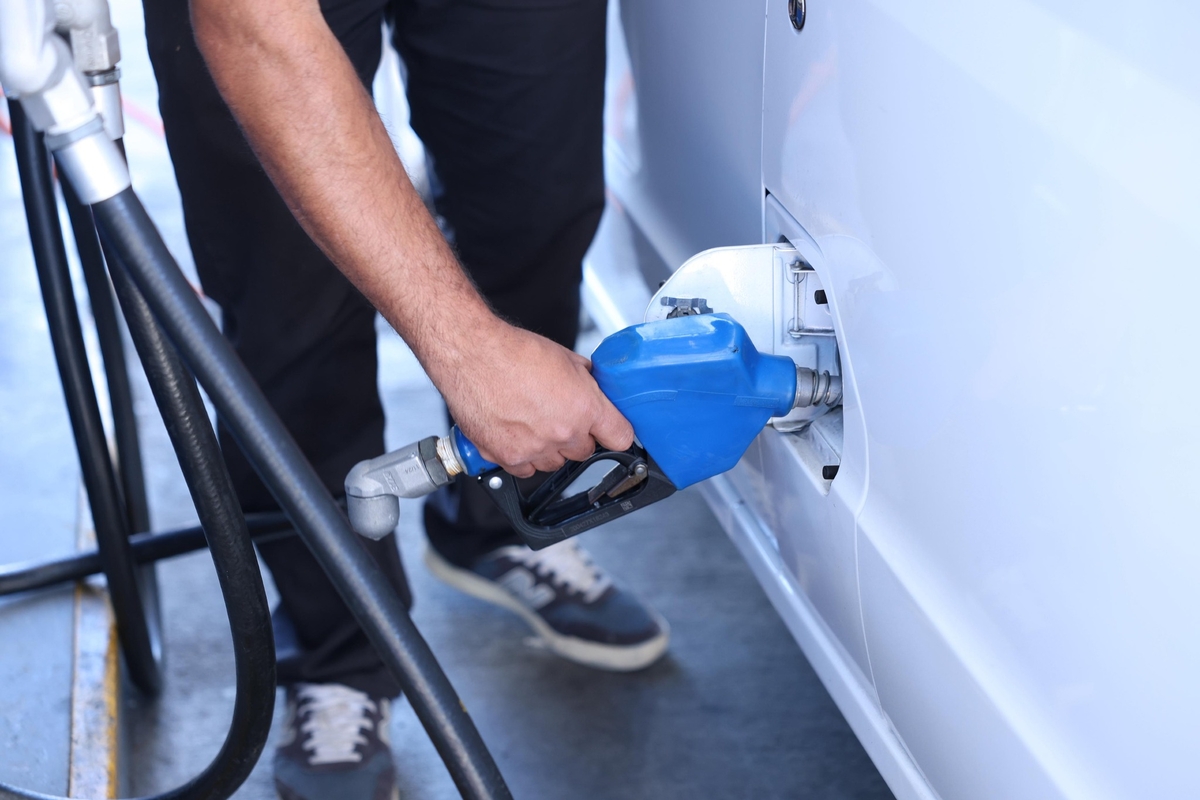Un hombre con casco blanco vierte líquido verde de un recipiente metálico a un cubo en una gasolinera, con un camión cisterna y surtidores de combustible al fondo