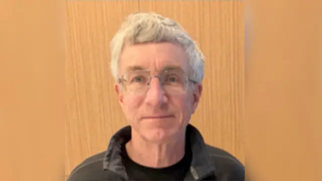 Una foto de William, que tiene el pelo blanco, gafas y una chaqueta con cuello.