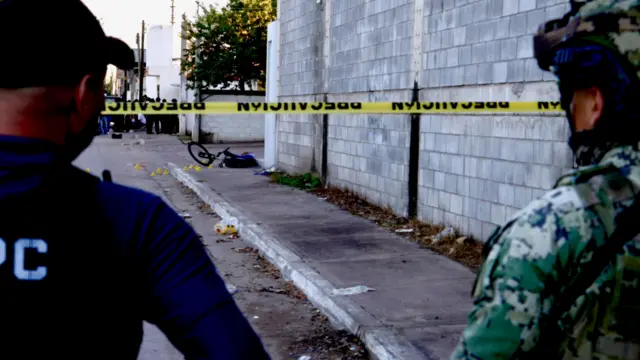 Un soldado y otro uniformado están detrás de una cinta de seguridad. A lo lejos, en la acera, un niño yace muerto junto a su bicicleta.
