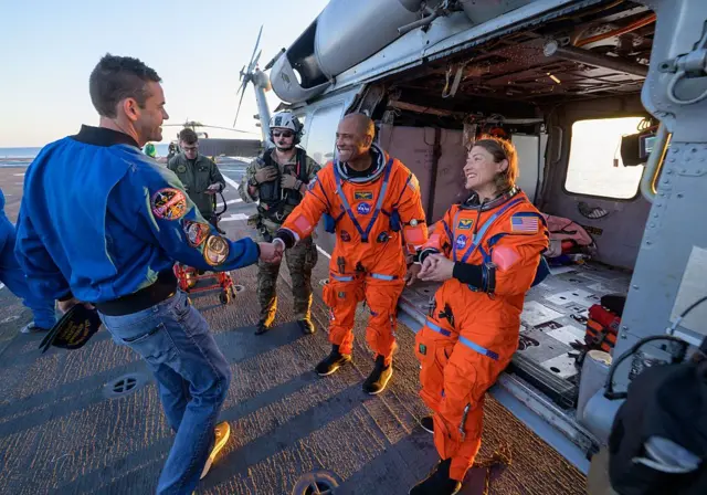 El astronauta Victor Glover, piloto de Artemis II (izq.) y la astronauta de la NASA Christina Koch, especialista en misiones Artemis II, salen de un helicóptero y conversan con el administrador de la NASA Jared Isaacman, a bordo del barco de rescate John P. Murtha