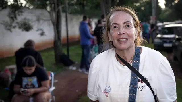 Zena, una señora mayor, vestida con una camisa blanca, cabello rubio y aros en las orejas y nariz.