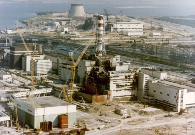 Vista de la central nuclear de Chernóbil el 29 de abril de 1986, tres días después de la explosión. El edificio que alberga el reactor número 4 presenta graves daños, con escombros ennegrecidos bajo una chimenea roja y blanca.