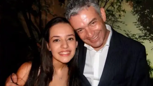 Victoria, vestida con un vestido negro, y su padre, que lleva una elegante chaqueta negra y una camisa blanca, sonríen a la cámara.