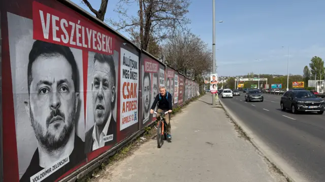 Un hombre pasa en bicicleta junto a un muro en una autopista de Hungría, empapelado con retratos de estilo cine negro de Volodymyr Zelensky y Péter Magyar.