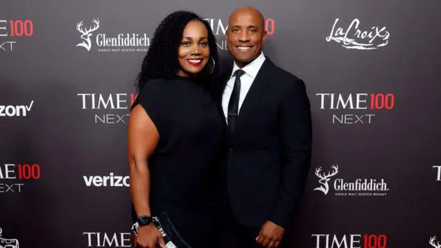Victor Glover está parado a la derecha, vestido en traje oscuro, camisa blanca y corbata delgada. Lo acompaña su esposa Dionna, con un vestido negro sencillo y agarra un bolso en su mano derecha. Ambos sonríen a cámara frente a una valla con los logotipos de la compañías patrocinadoras del evento