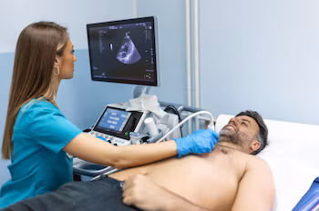 Una profesional médica en uniforme azul realiza un ultrasonido cardíaco a un hombre barbudo acostado, mientras un monitor muestra la imagen de un corazón