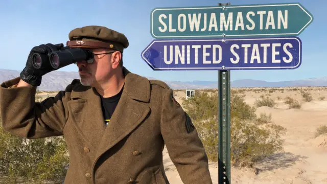 Un hombre uniformado con un abrigo y boina de color marrón, mira a la distancia a través de binoculares. Atrás hay dos avisos. Uno apunta hacia la izquierda y lee "Estados Unidos", el otro apunta hacia la derecha y lee "Slowjamastan". El paisaje en el fondo es desértico.