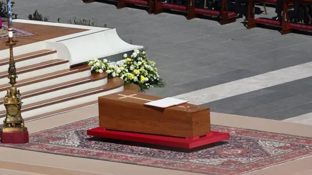 El féretro de madera, con una cruz dorada en la parte superior, está cerrado y yace sobre una plataforma roja. Debajo hay una alfombra de varios colores, entre ellos destaca el rojo, el azul y el beige.