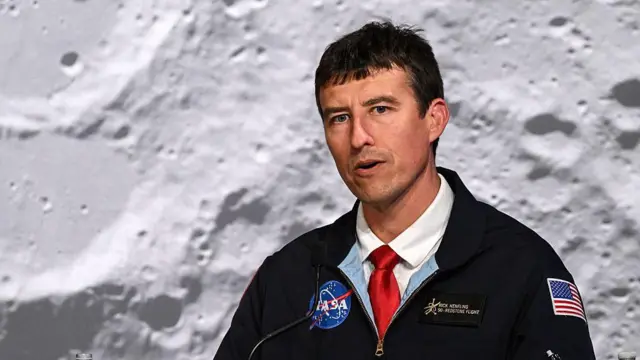 Un hombre de mediana edad, con una chaqueta negra de la NASA y una corbata roja, habla mirando ligeramente fuera de cámara, con una enorme imagen de la superficie lunar como fondo.