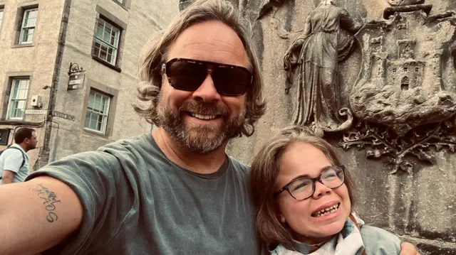 La imagen muestra a Darren y Sophia Scott en una selfie. Él tiene el cabello rubio, algo largo, y barba; lleva gafas de sol y una camiseta gris. Tiene el brazo alrededor de Sophia, quien lleva gafas y una bufanda. Ambos están sonriendo.Detrás de ellos se aprecia un monumento o escultura de piedra desgastada, con figuras talladas y lo que parece ser una cresta o un escudo de armas.