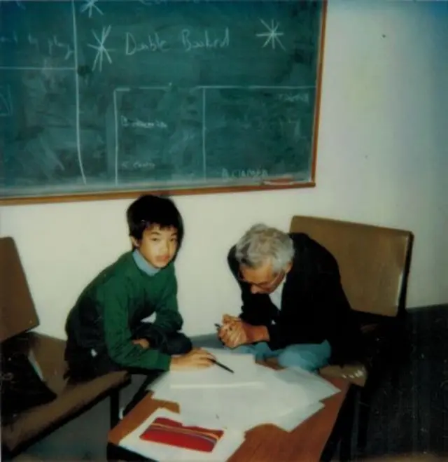 Terence Tao ve a la cámara mientras sostiene un bolígrafo. Se apoya en una mesa donde hay varias hojas. Al lado está Paul Erdős viendo los papeles