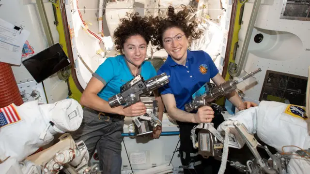 Las astronautas Jessica Meir y Christina Koch flotan dentro de la estación espacial, con sus cabellos elevados por la ingravidez. Visten playeras azules. Cada una sostiene una herramienta en la forma de taladro que sirve para ajustar los trajes espaciales