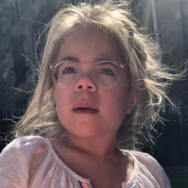 Sophia mira ligeramente fuera de cámara. Tiene el cabello largo y rubio recogido hacia atrás, y lleva unas gafas grandes de montura rosa y una blusa blanca de verano.