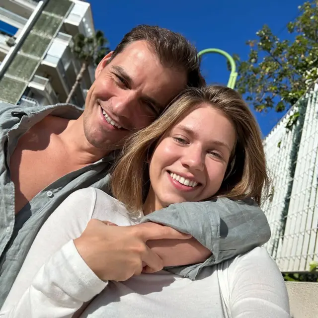 Mikhail y Kseniia abrazados mientras se toman un selfi sonriendo bajo un cielo azul