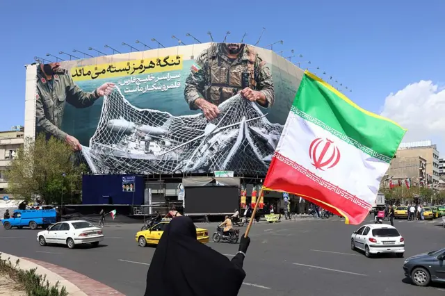 Una mujer con un hijab negro sostiene la bandera nacional de Irán mientras está de pie cerca de una valla publicitaria con la frase "El estrecho de Ormuz permanece cerrado" en la plaza Enqelab de Teherán, el 5 de abril de 2026.