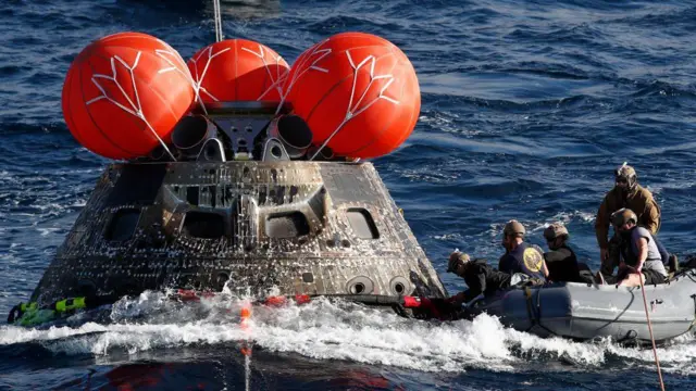 La cápsula Orión flota en el Pacífico mientras personal naval asegura un cable desde una balsa.