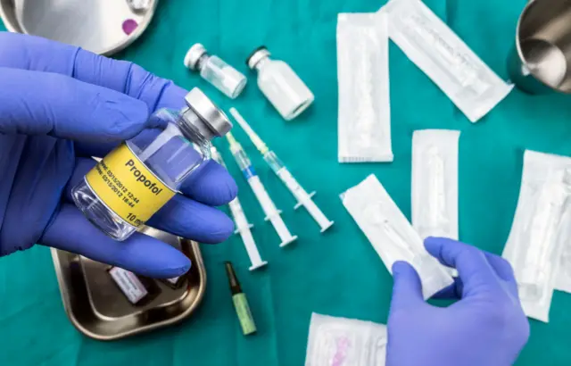 Un médico sostiene un frasco de propofol y de fondo una bandeja con inyecciones.