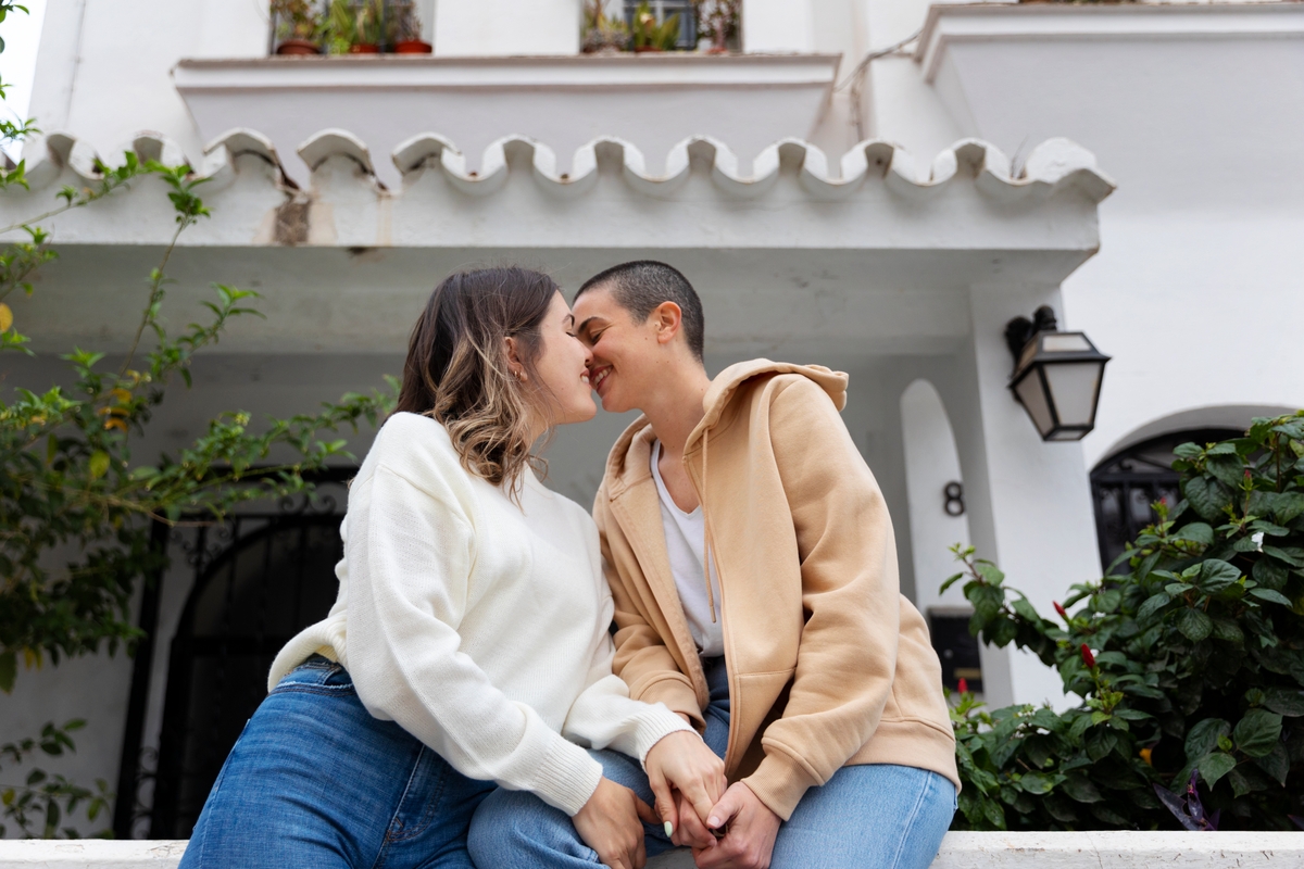 Dos mujeres se dan un beso tierno sentadas en un muro blanco, una con cabello ondulado y suéter blanco, la otra con cabello corto y sudadera beige