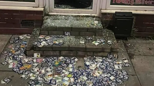 En la entrada de una tienda se ven cientos de cartas Pokémon esparcidas por el suelo, mezcladas con cristales rotos de la puerta principal.