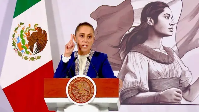 La presidenta mexicana, Claudia Sheinbaum, se encuentra en un atril frente a una bandera mexicana y un mural que representa a una mujer mexicana durante su conferencia de prensa habitual. Lleva una chaqueta azul con bordados dorados en las solapas. Lleva el cabello recogido y gesticula mientras habla.